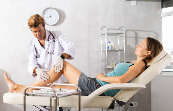 A male doctor checking the left leg of a young woman lying in a feet elevated position on an adjustable hospital bed.